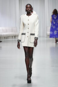 Model walks the runway in a cream textured coat featuring gold buttons and black embroidered trim on pockets and cuffs.