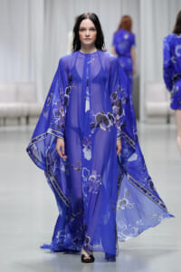Model walks the runway in a sheer cobalt blue kimono dress with large white and pale blue floral print and wide flowing sleeves, paired with black sandals.