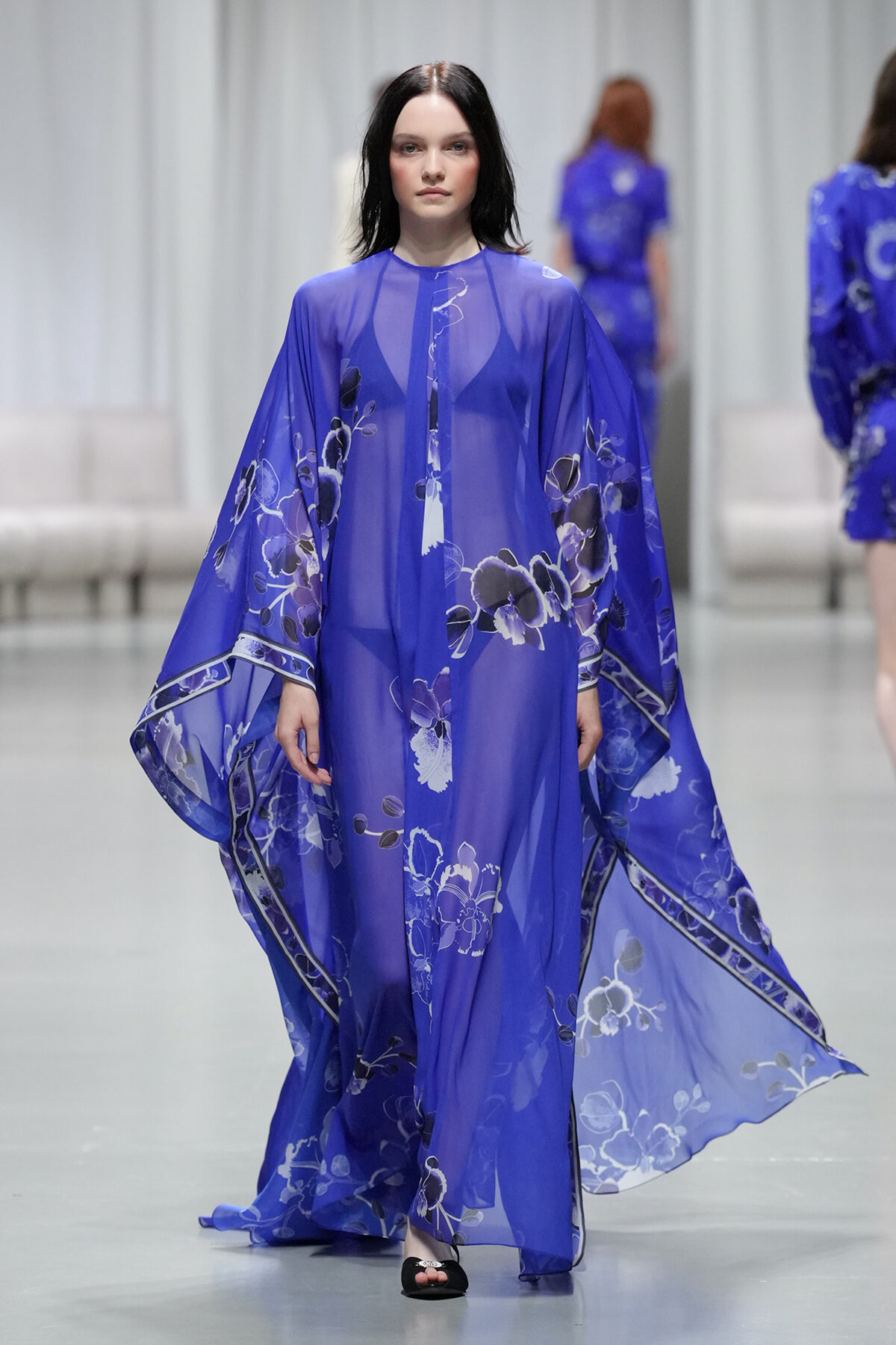 Model walks the runway in a sheer cobalt blue kimono dress with large white and pale blue floral print and wide flowing sleeves, paired with black sandals.