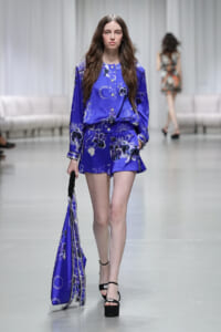 Model walking a fashion runway in a cobalt blue floral-print blouse and matching shorts, carrying a matching blue scarf bag.
