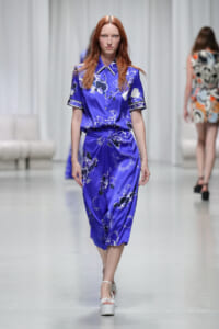 Female model walking the runway in a cobalt blue floral-print satin dress with short sleeves and a tied waist, white platform sandals visible.