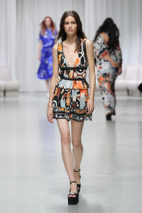 Model walking a fashion runway in a sleeveless multicolored abstract-print romper with a peplum waist and black belt, wearing black platform sandals.