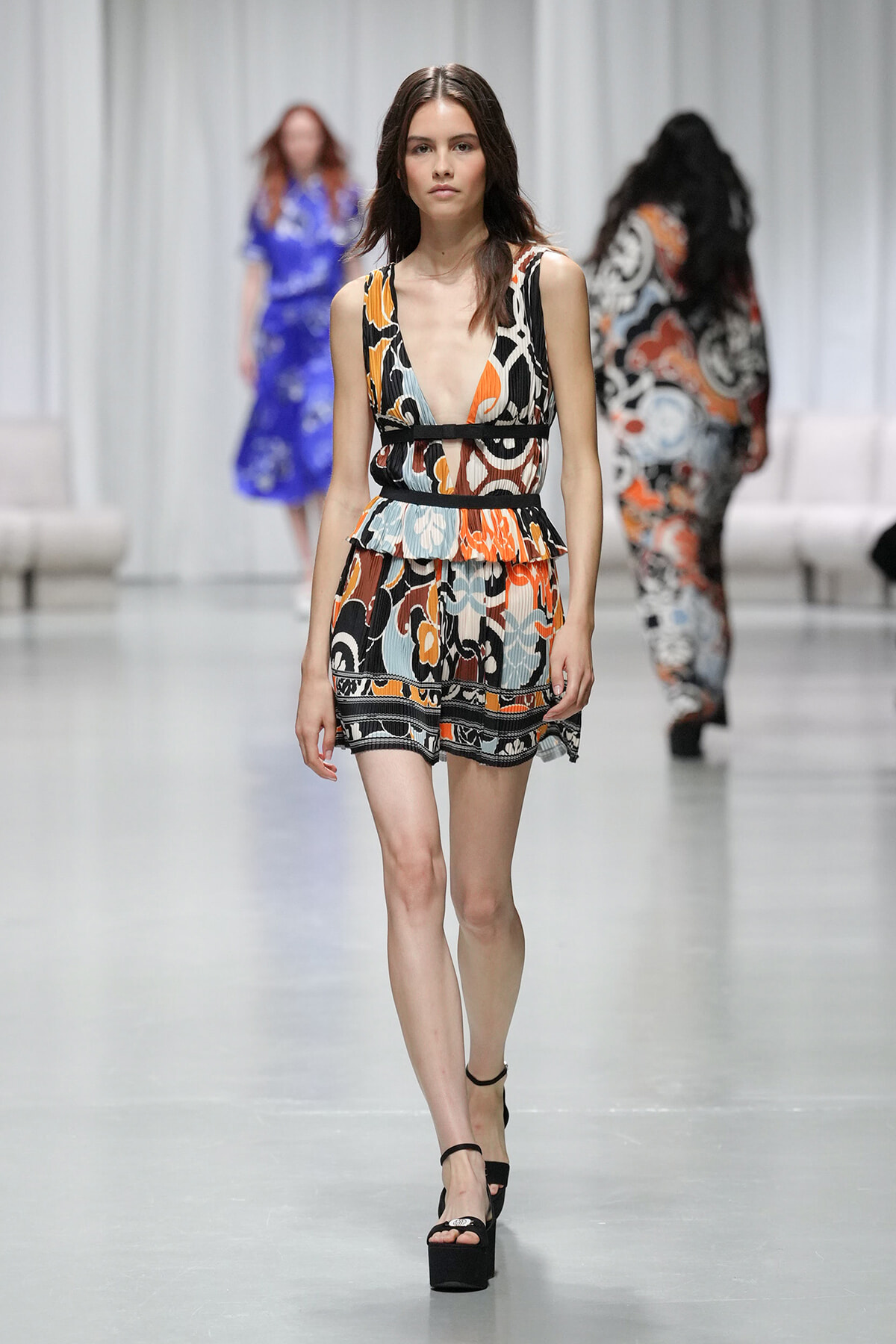 Model walking a fashion runway in a sleeveless multicolored abstract-print romper with a peplum waist and black belt, wearing black platform sandals.