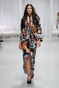 Model walks the runway in a long abstract floral wrap dress in orange, black, blue, and white, tied at the waist with a black neck strap; black platform sandals.