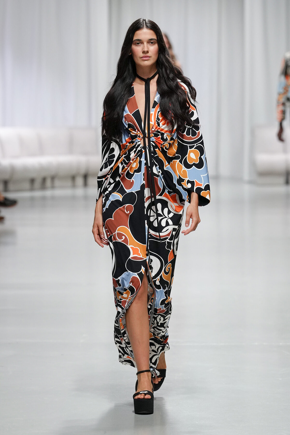 Model walks the runway in a long abstract floral wrap dress in orange, black, blue, and white, tied at the waist with a black neck strap; black platform sandals.
