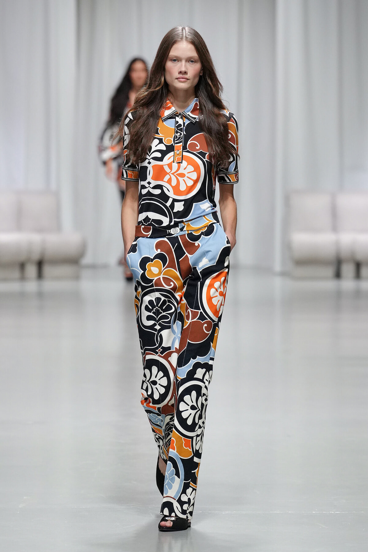 Model walks a fashion runway wearing a bold orange, black, white, and blue abstract floral print matching blouse and wide-leg pants with hands in pockets.