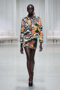 Model walks a runway wearing a bold patterned orange, black, white and blue blazer with matching mini skirt and platform heels.