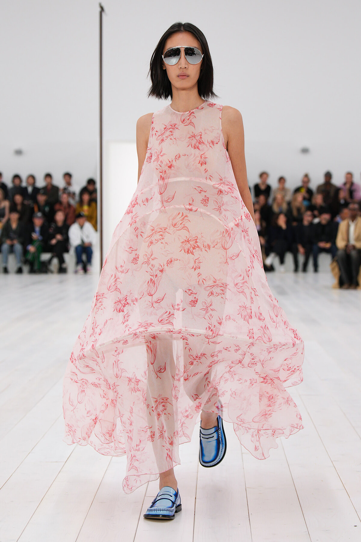 Female model walking a runway in a sheer pink floral dress with oversized sunglasses and blue loafers; audience seated in the background.