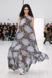 Female model walks a fashion runway in a sleeveless maxi dress with large lavender floral print on dark background.