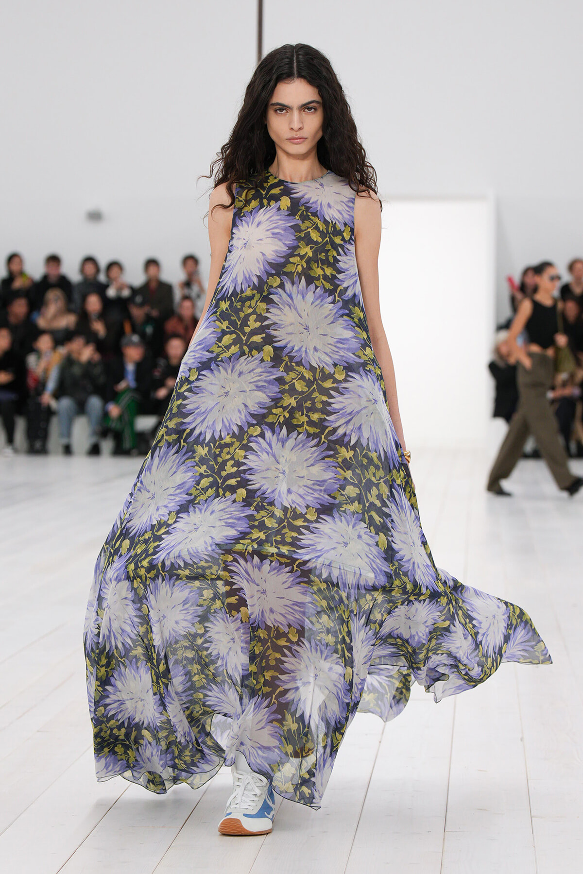Female model walks a fashion runway in a sleeveless maxi dress with large lavender floral print on dark background.