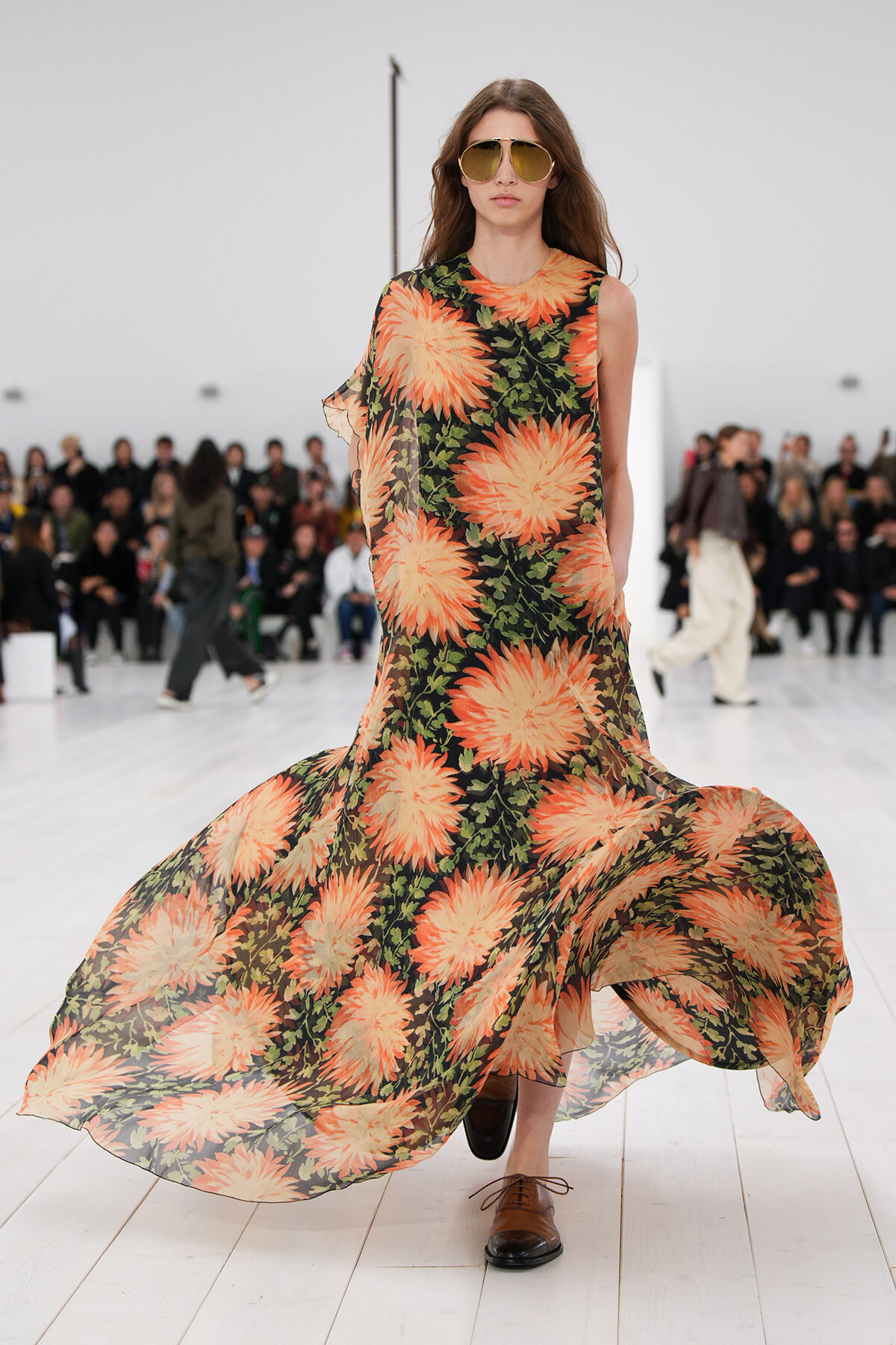 Model walking the runway in a long black dress with large orange floral print and sunglasses.
