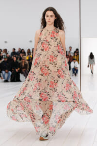 Model walks a fashion show in a flowing sleeveless beige dress with pink and red rose print, on a white runway.