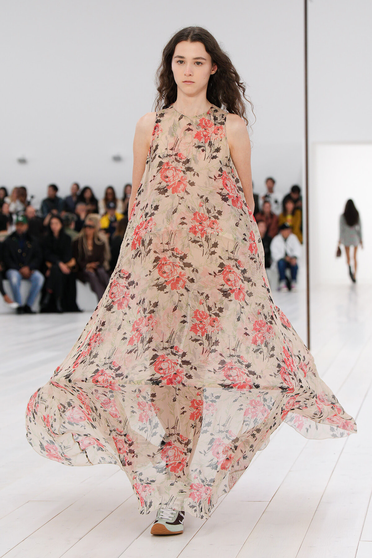 Model walks a fashion show in a flowing sleeveless beige dress with pink and red rose print, on a white runway.