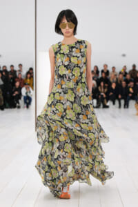 Model walking a runway in a sleeveless, floor-length floral dress with green, yellow, and white flowers on black; orange sneakers and large aviator sunglasses, audience in background.