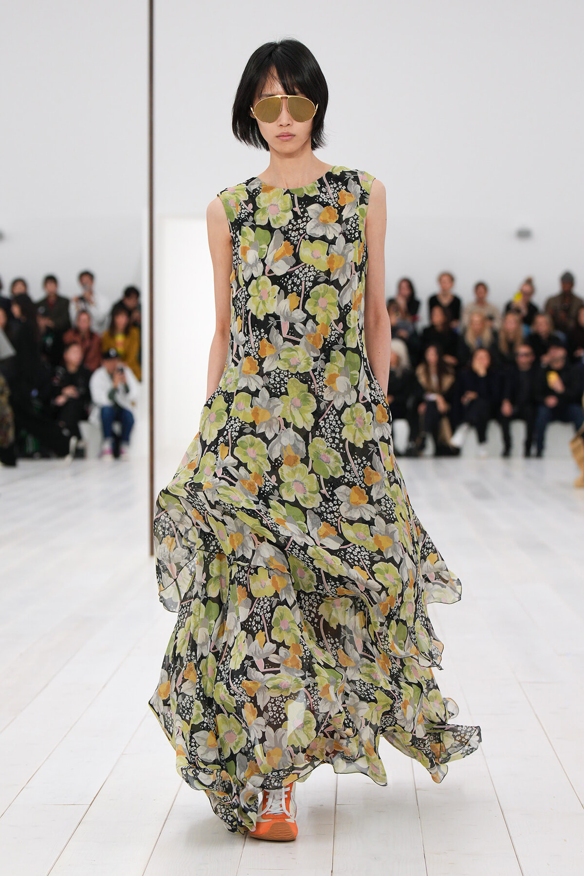 Model walking a runway in a sleeveless, floor-length floral dress with green, yellow, and white flowers on black; orange sneakers and large aviator sunglasses, audience in background.