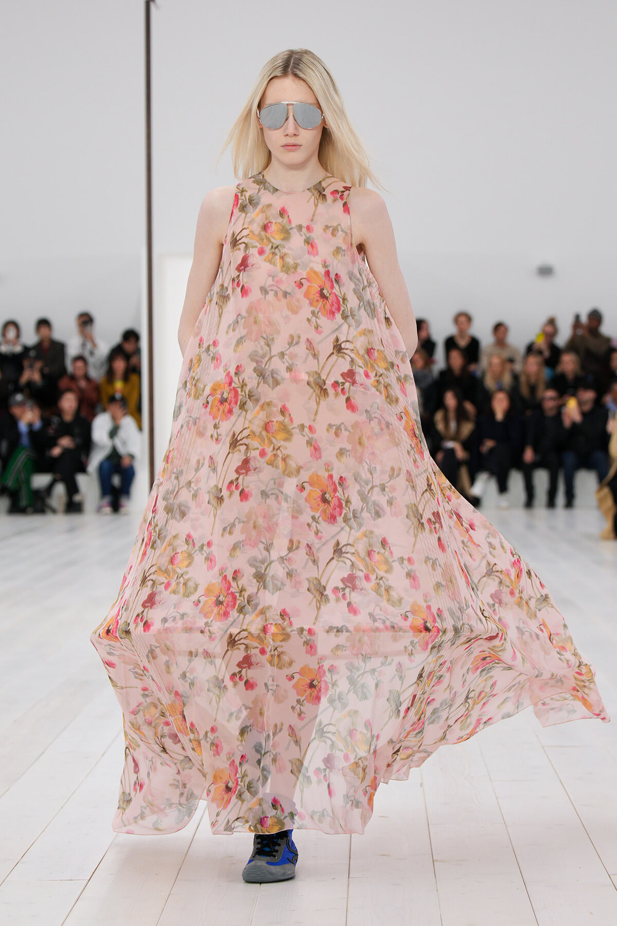 Model walking a fashion runway in a flowing pink floral maxi dress with mirrored sunglasses, audience in the background