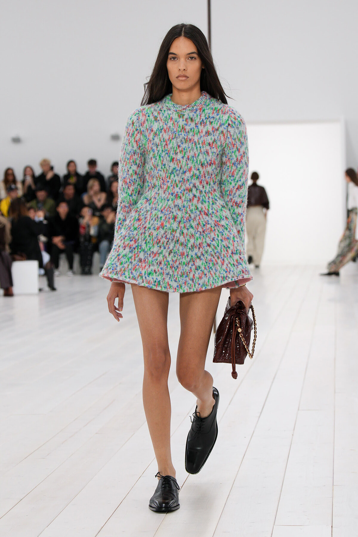 Model walking the runway in a short, speckled knit dress with long sleeves; she holds a brown handbag with a gold chain and wears black lace-up shoes.