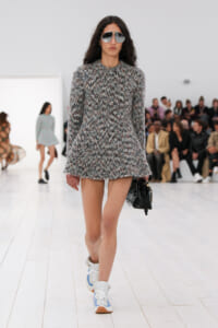 Model walks a fashion runway in a gray speckled mini dress with matching sunglasses and white sneakers, carrying a black bag; audience in background.