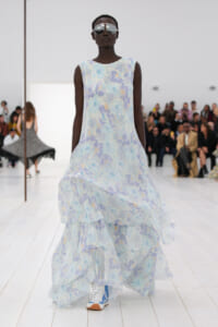 Model walks the runway in a light blue floral sleeveless dress with a tiered, flowing hem and oversized sunglasses; seated audience in background.
