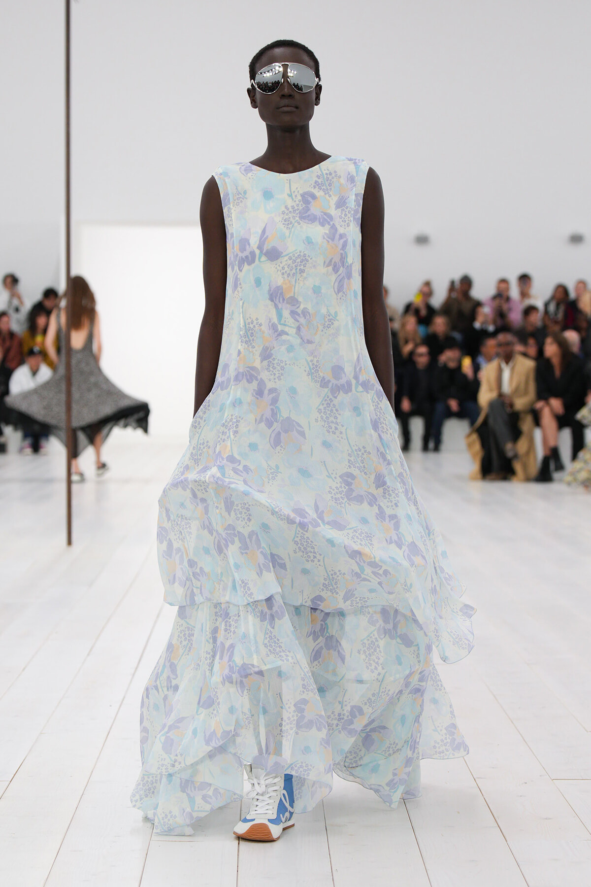 Model walks the runway in a light blue floral sleeveless dress with a tiered, flowing hem and oversized sunglasses; seated audience in background.