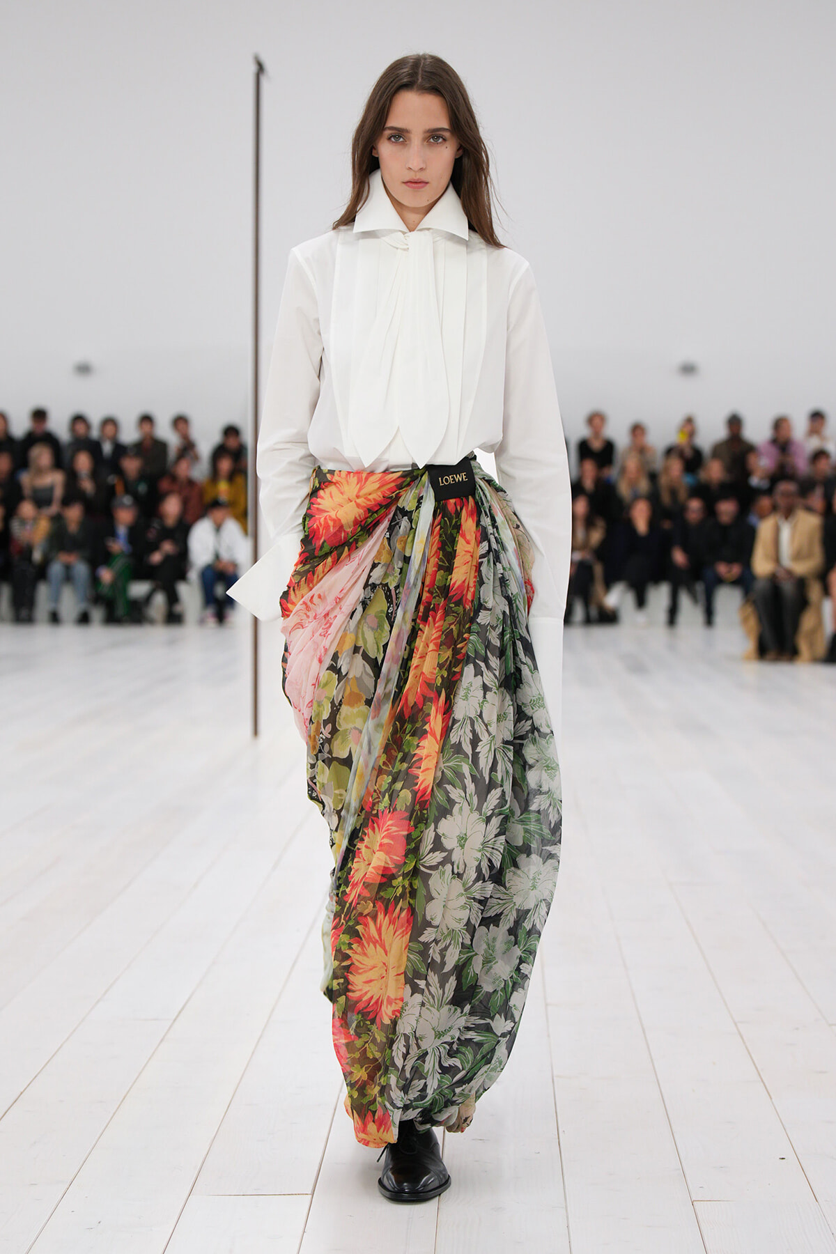 Model walking a runway in a white blouse and a vibrant floral wrap skirt tied with a Loewe belt; audience seated in the background.