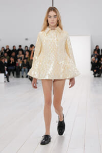 Model walking the runway in a pale, metallic cream cape-like coat with a flared A-line hem and wide collar.