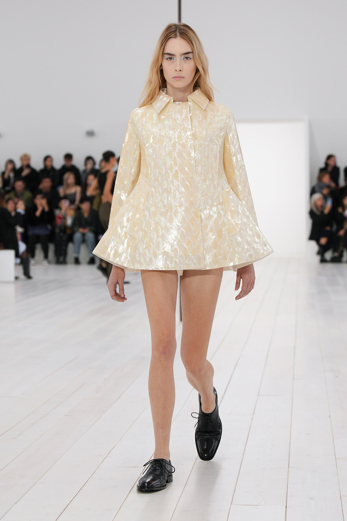 Model walking the runway in a pale, metallic cream cape-like coat with a flared A-line hem and wide collar.