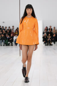 Model walks the runway wearing an orange sequined long-sleeve mini dress with a flared skirt and burgundy lace-up shoes, audience in the background.