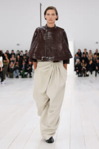 Model walking a runway in a dark brown crocodile-embossed cape jacket and oversized beige draped pants, audience in background