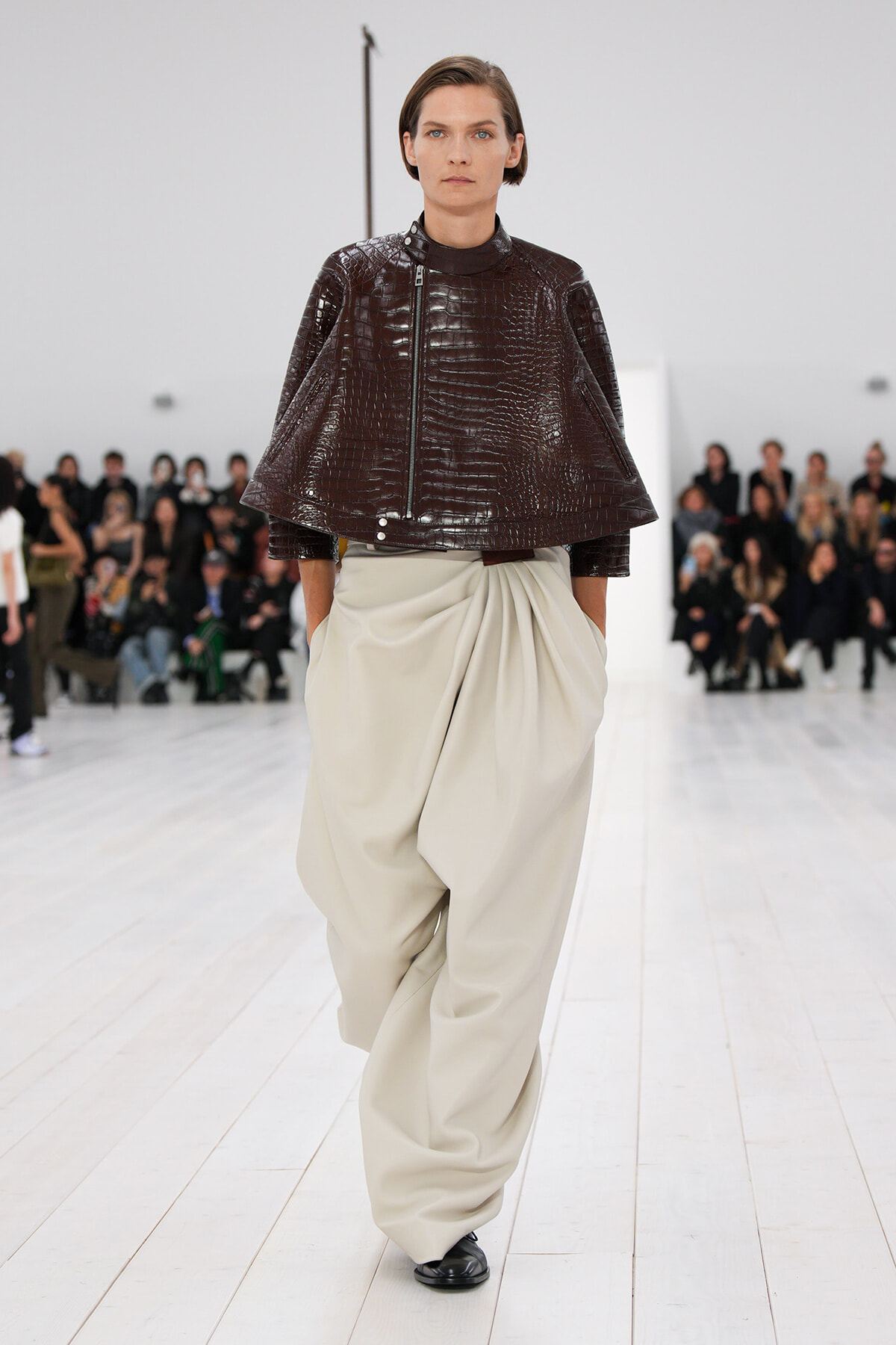 Model walking a runway in a dark brown crocodile-embossed cape jacket and oversized beige draped pants, audience in background