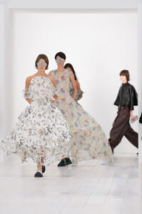 Models walk the runway in flowing floral gowns with oversized sunglasses in a bright white studio setting, mid-stride.
