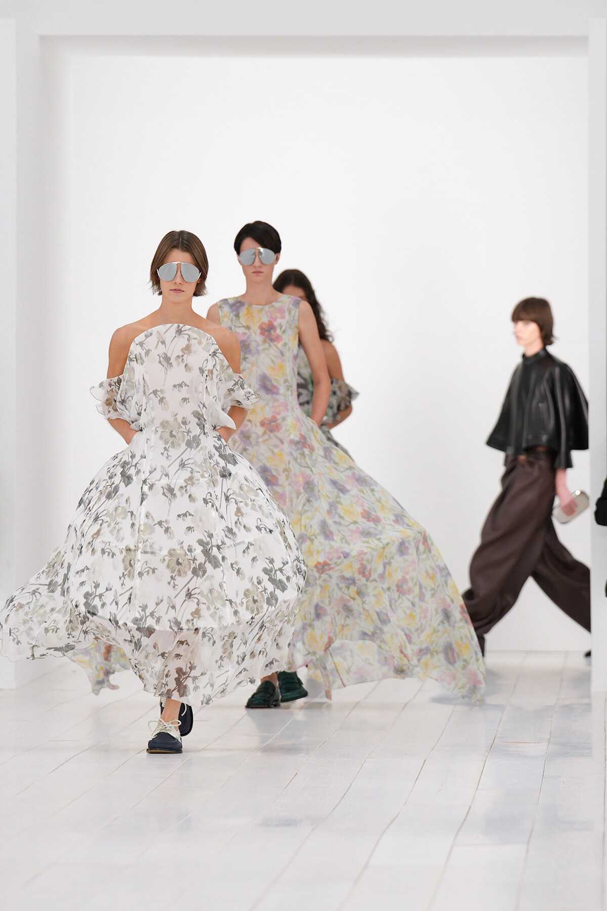 Models walk the runway in flowing floral gowns with oversized sunglasses in a bright white studio setting, mid-stride.