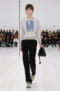 Model walks a fashion show wearing a light gray patterned sweater with a blue graphic, black trousers, and floral sneakers, carrying a black handbag.