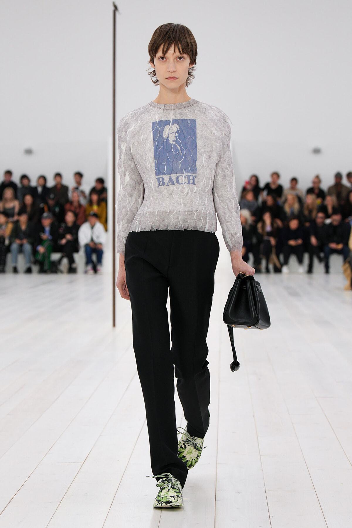 Model walks a fashion show wearing a light gray patterned sweater with a blue graphic, black trousers, and floral sneakers, carrying a black handbag.