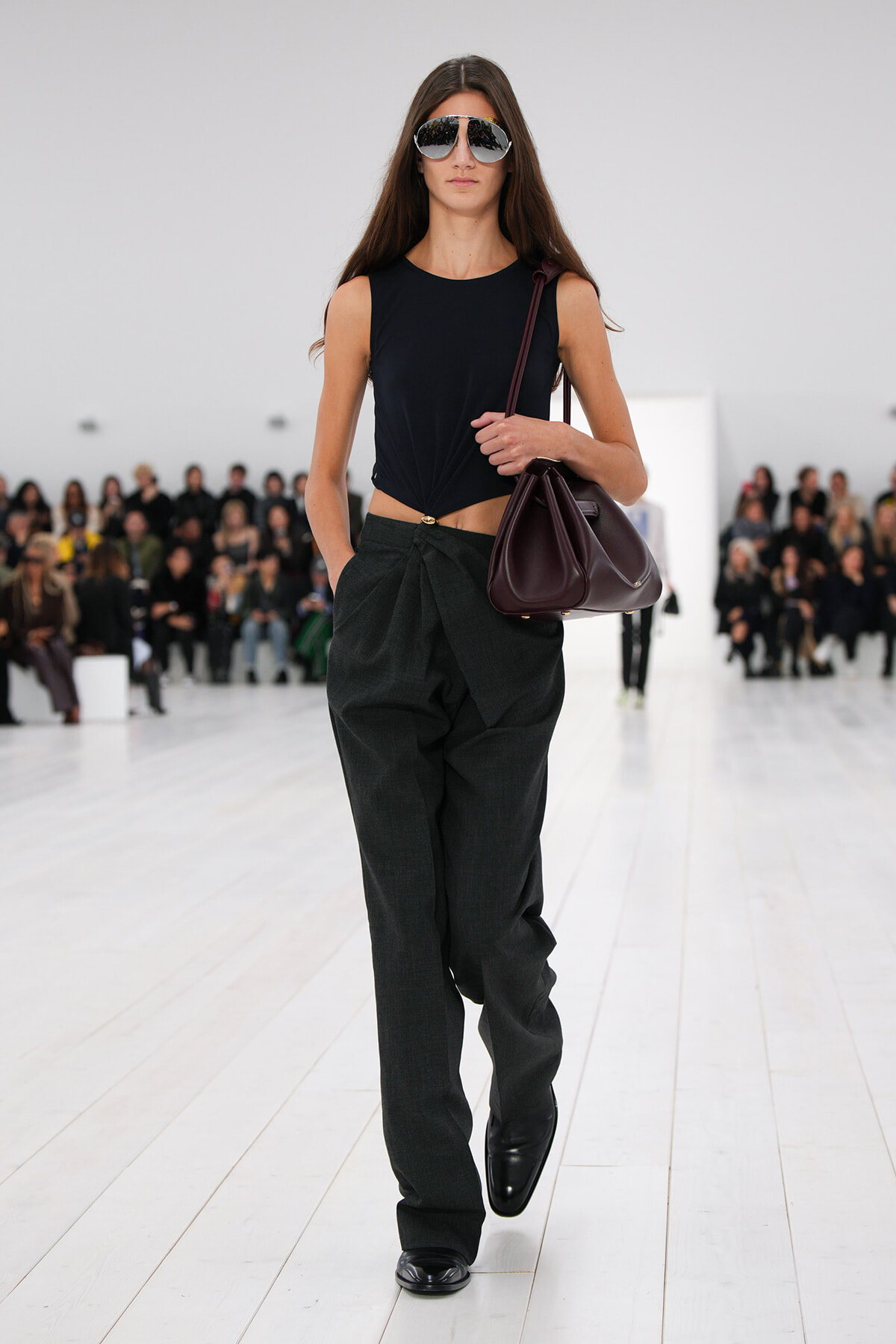 Model walking a runway in a sleeveless black knotted top, dark wide-leg pants, and black shoes, carrying a burgundy handbag and wearing mirrored sunglasses.