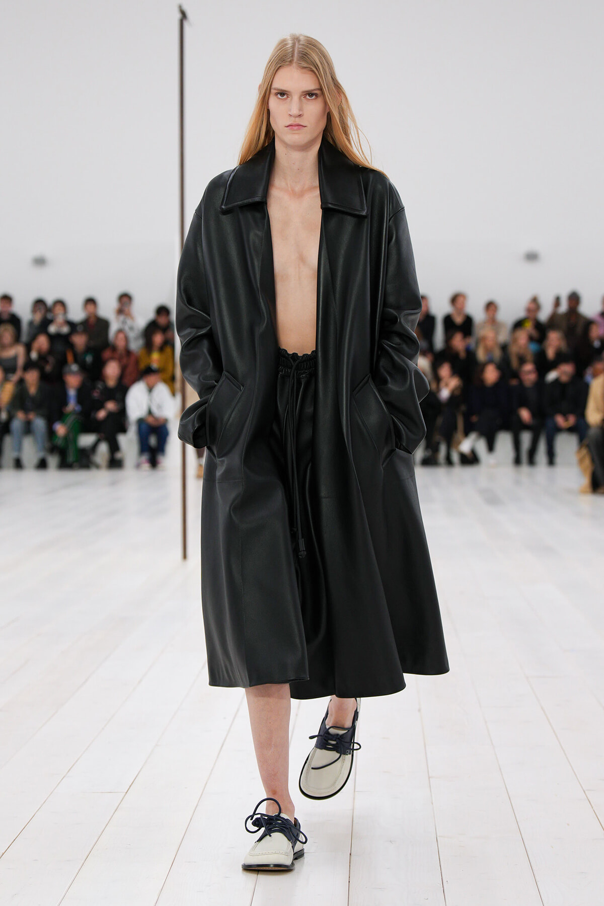 Model walking a fashion show in a long black leather coat open at the chest, with black drawstring pants and white shoes; audience sits in the background.