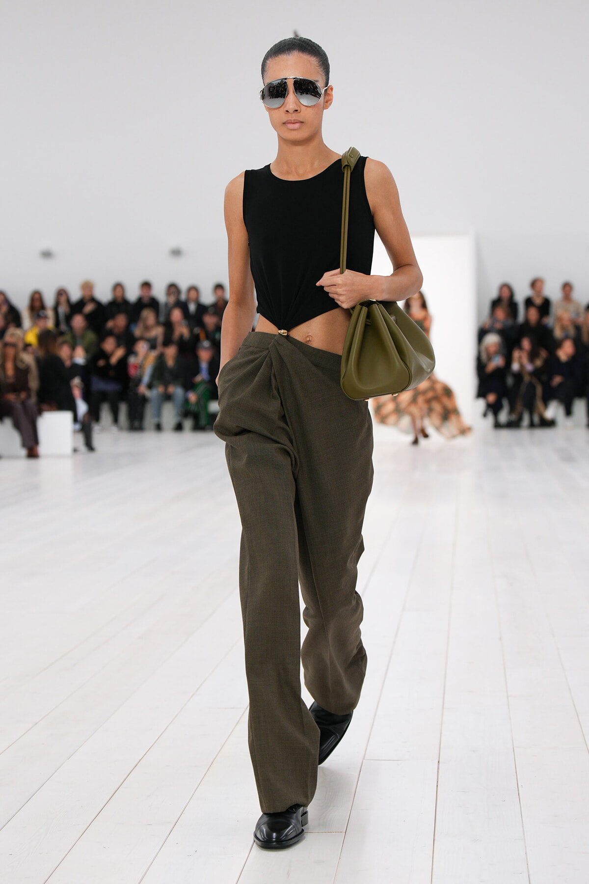 Model on a runway wearing a black sleeveless top knotted at the waist, olive wide pants, black shoes, and an olive shoulder bag with sunglasses.