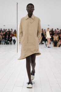 Runway model in a beige coat with hands in pockets, walking on a white catwalk as the audience watches.