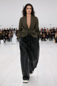 Model walking down a runway in an olive blazer with a deep V and oversized black draped trousers, white sneakers, audience in the background.