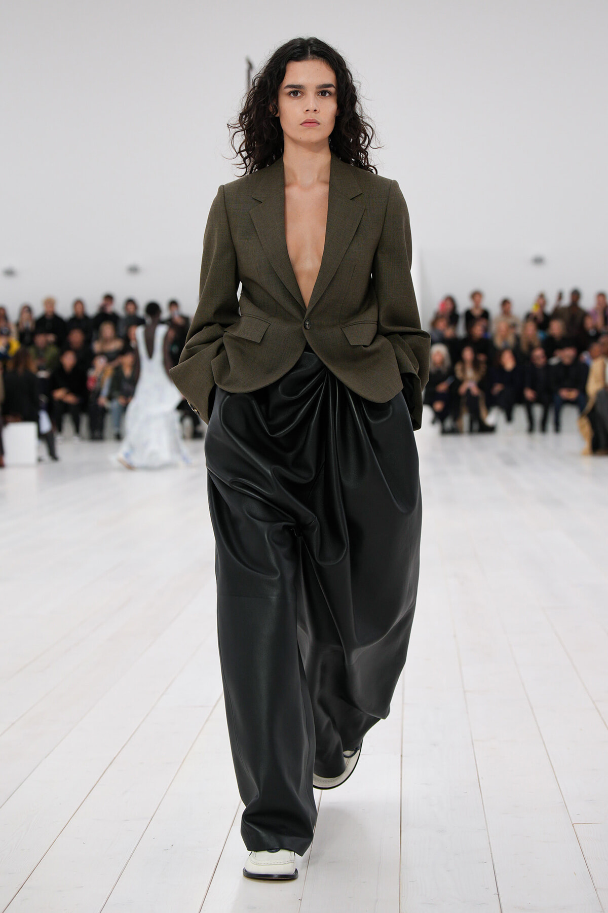 Model walking down a runway in an olive blazer with a deep V and oversized black draped trousers, white sneakers, audience in the background.