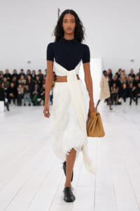 Model walks down a white runway in a navy polo top, white wrap skirt, and black shoes, carrying a tan handbag.