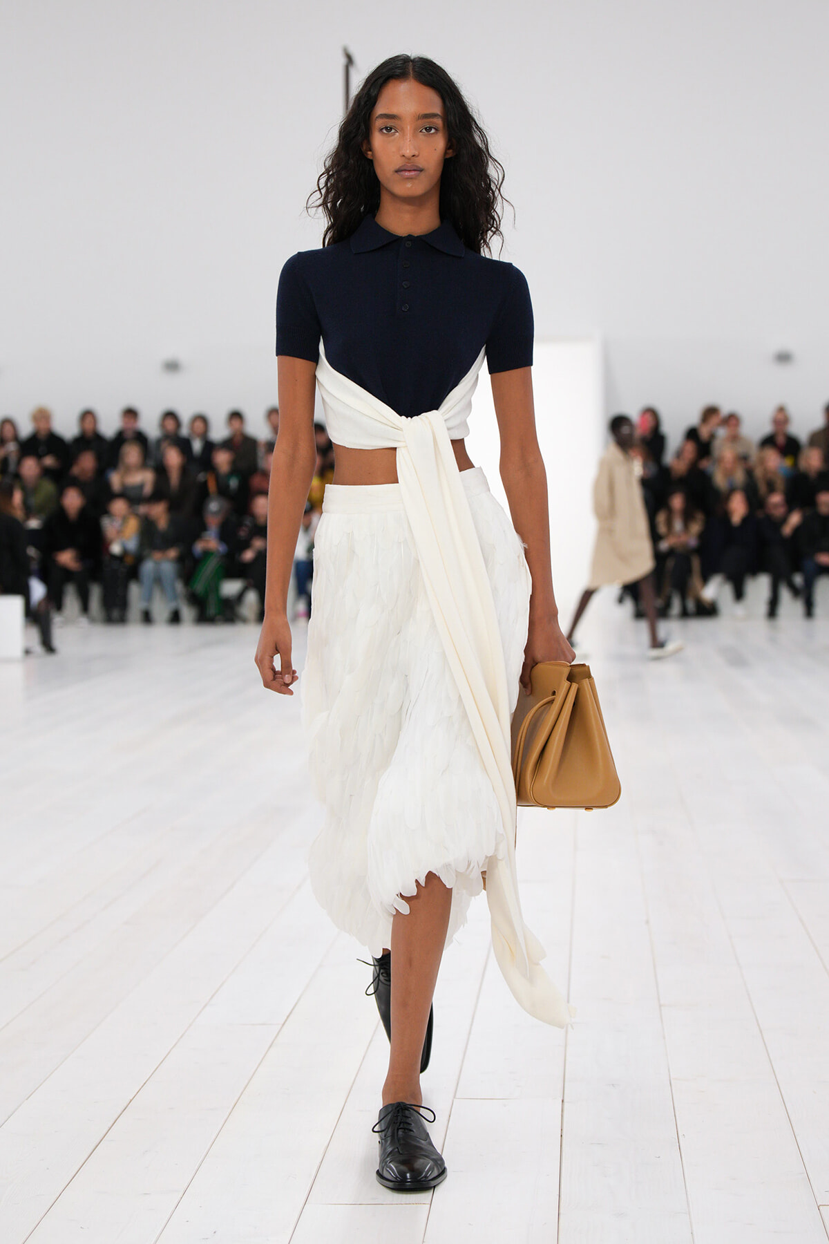 Model walks down a white runway in a navy polo top, white wrap skirt, and black shoes, carrying a tan handbag.
