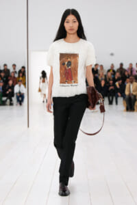 Model on a fashion runway wearing a white feathered top with a printed artwork, black trousers, and a brown handbag; audience in the background.