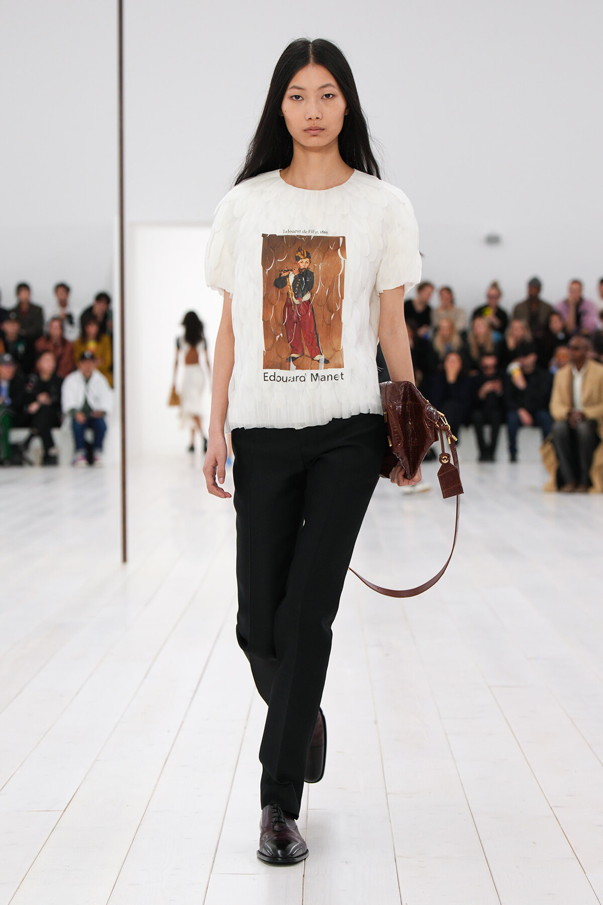 Model on a fashion runway wearing a white feathered top with a printed artwork, black trousers, and a brown handbag; audience in the background.