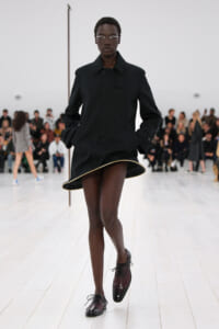 Runway model in a black structured coat dress walks toward the camera with hands in pockets, audience in the background.
