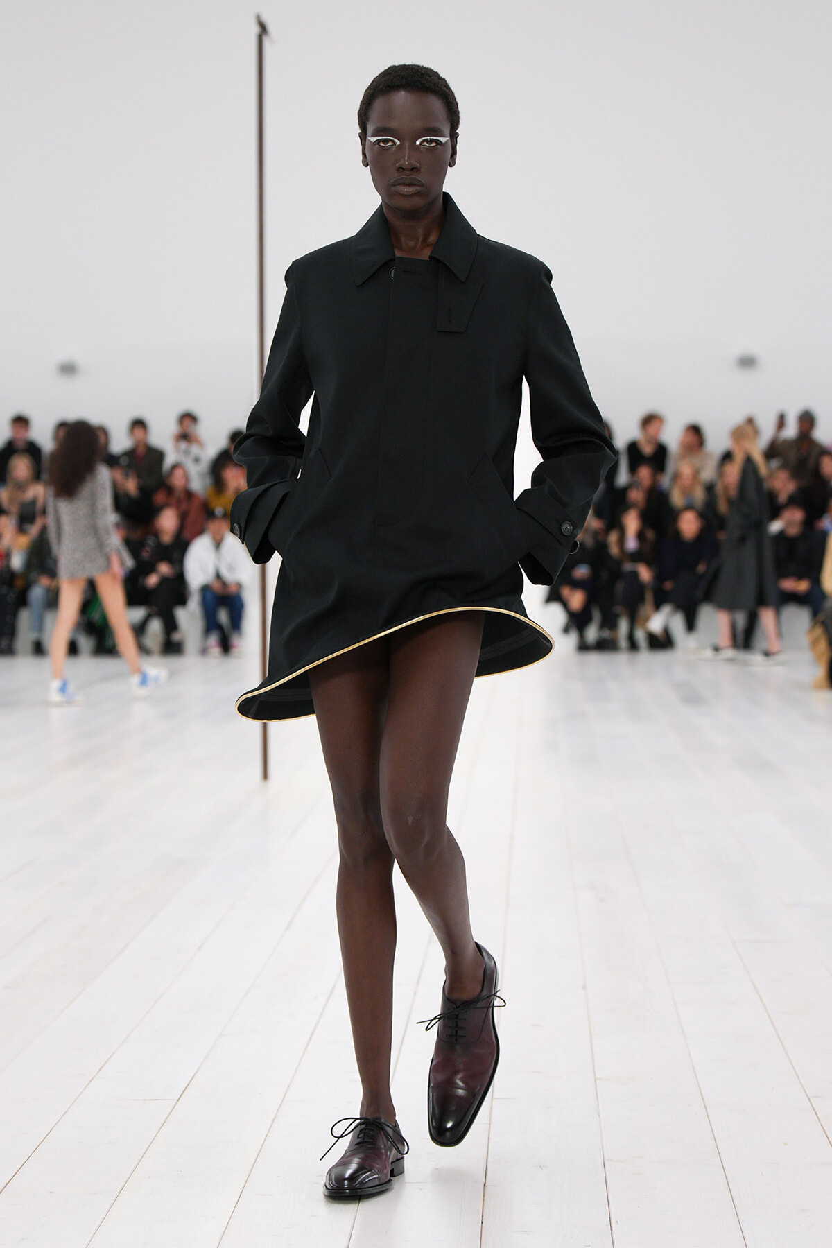 Runway model in a black structured coat dress walks toward the camera with hands in pockets, audience in the background.