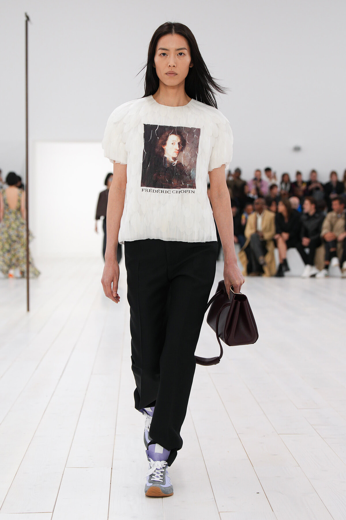 Model walking a fashion runway in a white feathered top with Frédéric Chopin print, black trousers, and purple sneakers, carrying a burgundy handbag