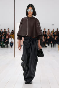Model walking a runway in a brown oversized leather cape jacket and wide black satin draped pants, accessorized with silver sunglasses and a black handbag