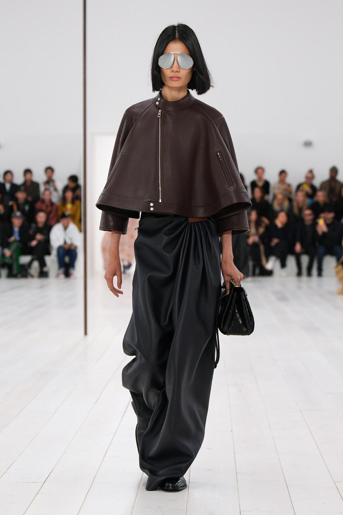 Model walking a runway in a brown oversized leather cape jacket and wide black satin draped pants, accessorized with silver sunglasses and a black handbag