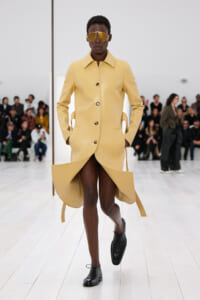 Model walking a fashion runway in a pale yellow, tailored coat with a dramatic curved hem, gold sunglasses, and black lace-up shoes.